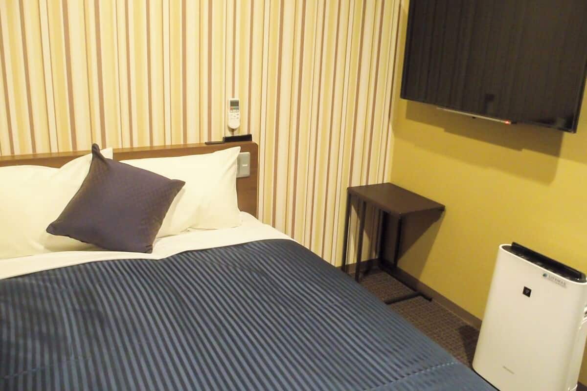 A hotel room with a bed, a bedside table, a TV mounted on the wall, and a white air conditioning unit.