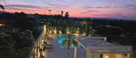 A rooftop pool area with lounge chairs and umbrellas, surrounded by greenery and a view of the landscape.