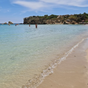 A beach with clear shallow waters, a sandy shore, and rocky cliffs in the background. - Villa Merilusi – Luxury Villa, Private Pool & Sea View (siracusa)
