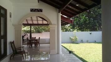 A covered patio with a table and chairs, a small garden area with grass and plants, and a white wall with a potted plant.
