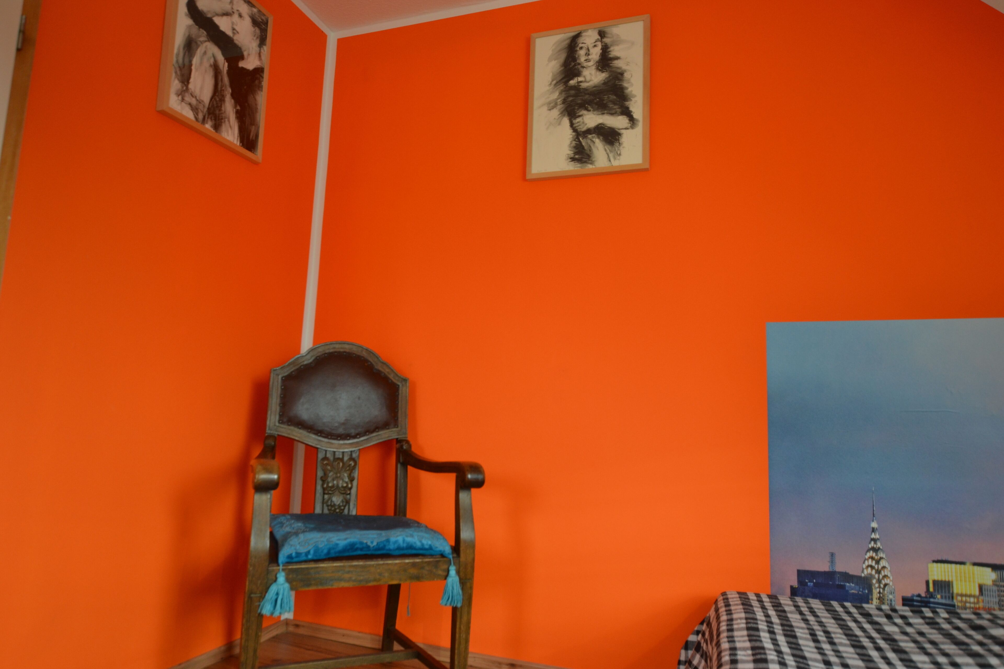 Une pièce aux murs orange, une chaise en bois avec un coussin bleu et un lit recouvert d’une couverture à carreaux.
