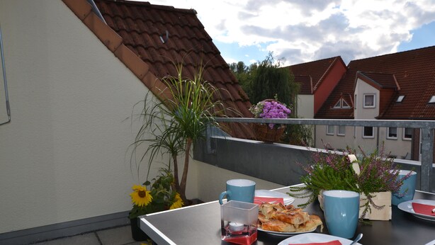 En balkong med ett bord dukat för två personer, med en blå mugg, tallrikar, servetter och ett litet ljus.