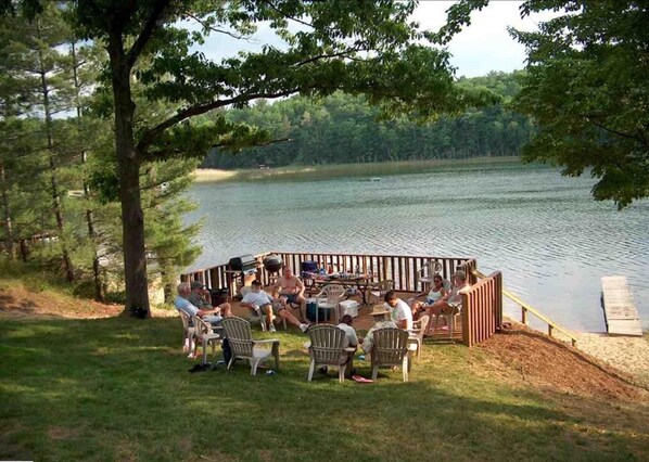 Relax on the lakeside deck with Adirondack chairs and a small table, surrounded by lush greenery.