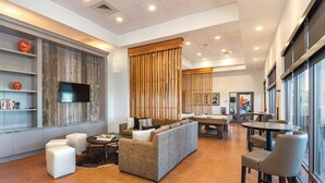 A modern living room with a sofa, armchairs, a coffee table, and a TV mounted on a wooden wall. There is a large window with blinds, a door leading to a gym, and a bookshelf with books and decorative items. - Gorgeous views of the rural surroundings! (Marble Falls)