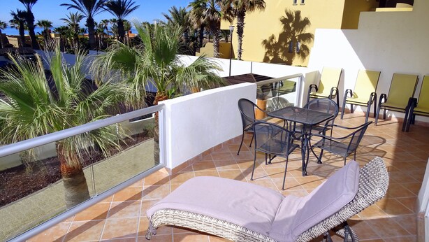 Ein Balkon mit einer Korbsitzlounge, einem Metalltisch und Stühlen. Palmen und Blick auf ein Gebäude mit gelber Fassade.