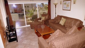 Ein Wohnzimmer mit einem braunen Sofa, einem Holztisch, einem Fernseher und einer Glastür, die auf einen Balkon mit Pflanzen führt.