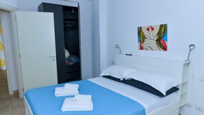 Ein ordentlich bezogenes Bett mit blauer Bettdecke und weißen Kissen, ein weißer Nachttisch und ein an der Wand hängendes Gemälde.