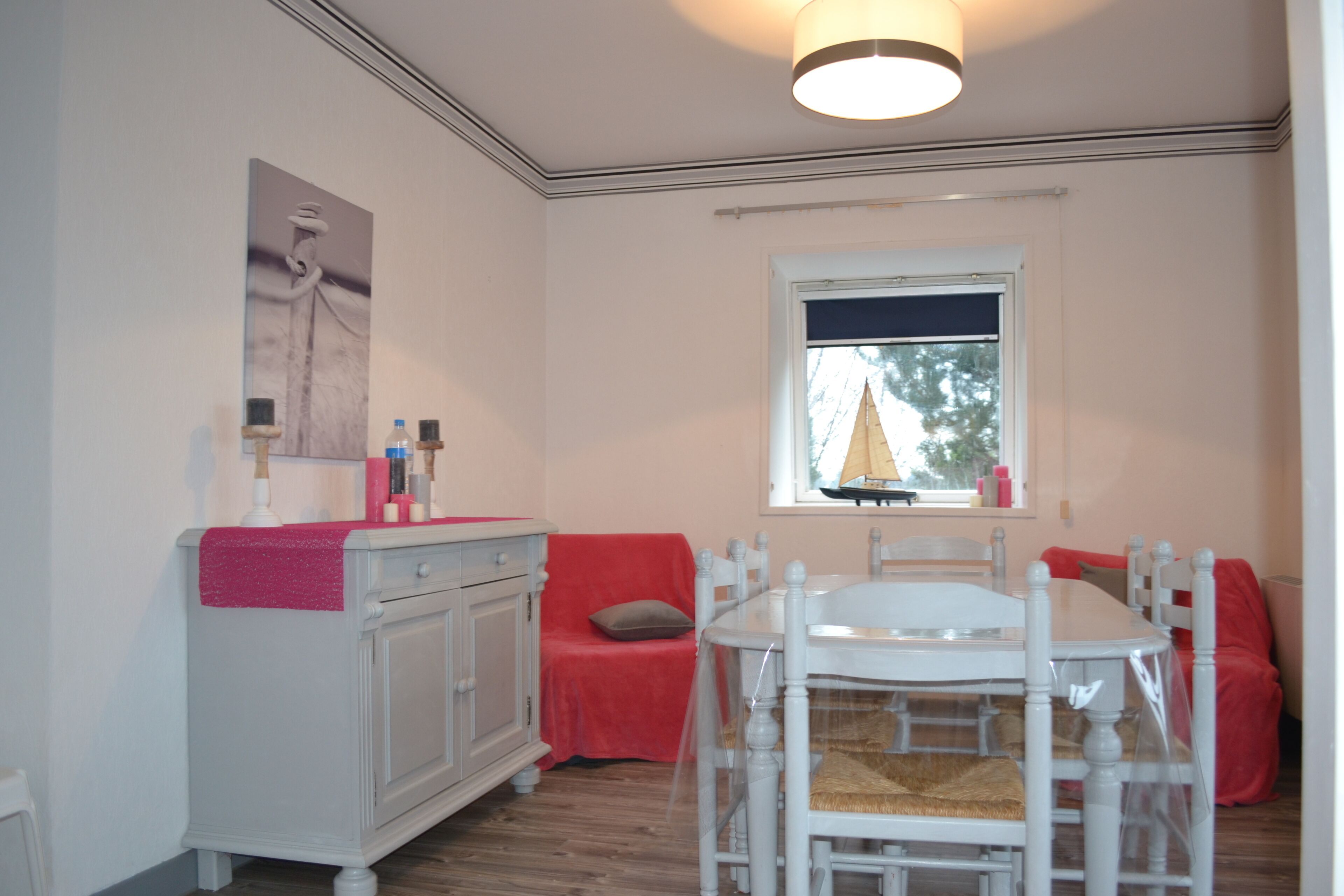 A dining area with a white table and chairs, a red sofa, a window with a view of trees, and a white cabinet with a pink towel.