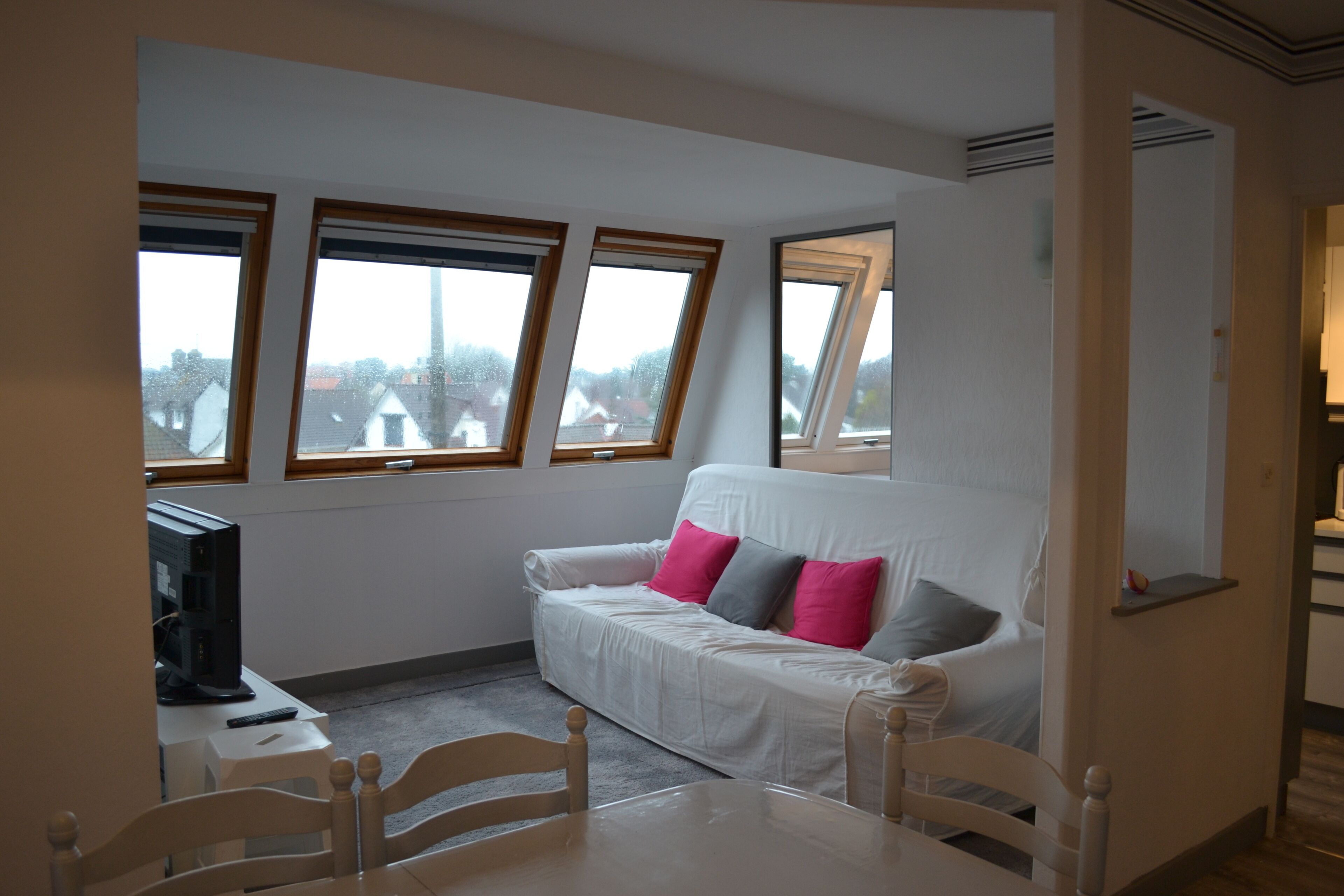 A room with a white sofa, a television on a small table, and three windows with wooden frames.