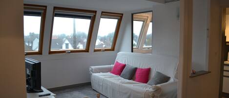 A room with a white sofa, a television on a small table, and three windows with wooden frames.