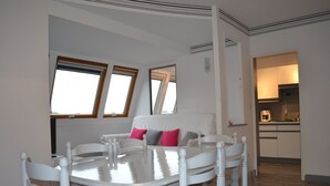 Zona pranzo con tavolo rotondo e sedie, divano con cuscini e cucina sullo sfondo.