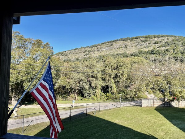 A view of a grassy area with a flagpole displaying an American flag, a metal fence, and a hilly landscape in the background.