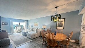 Uma sala de estar espaçosa com área de jantar, apresentando uma grande janela, um candelabro e vista para o exterior.