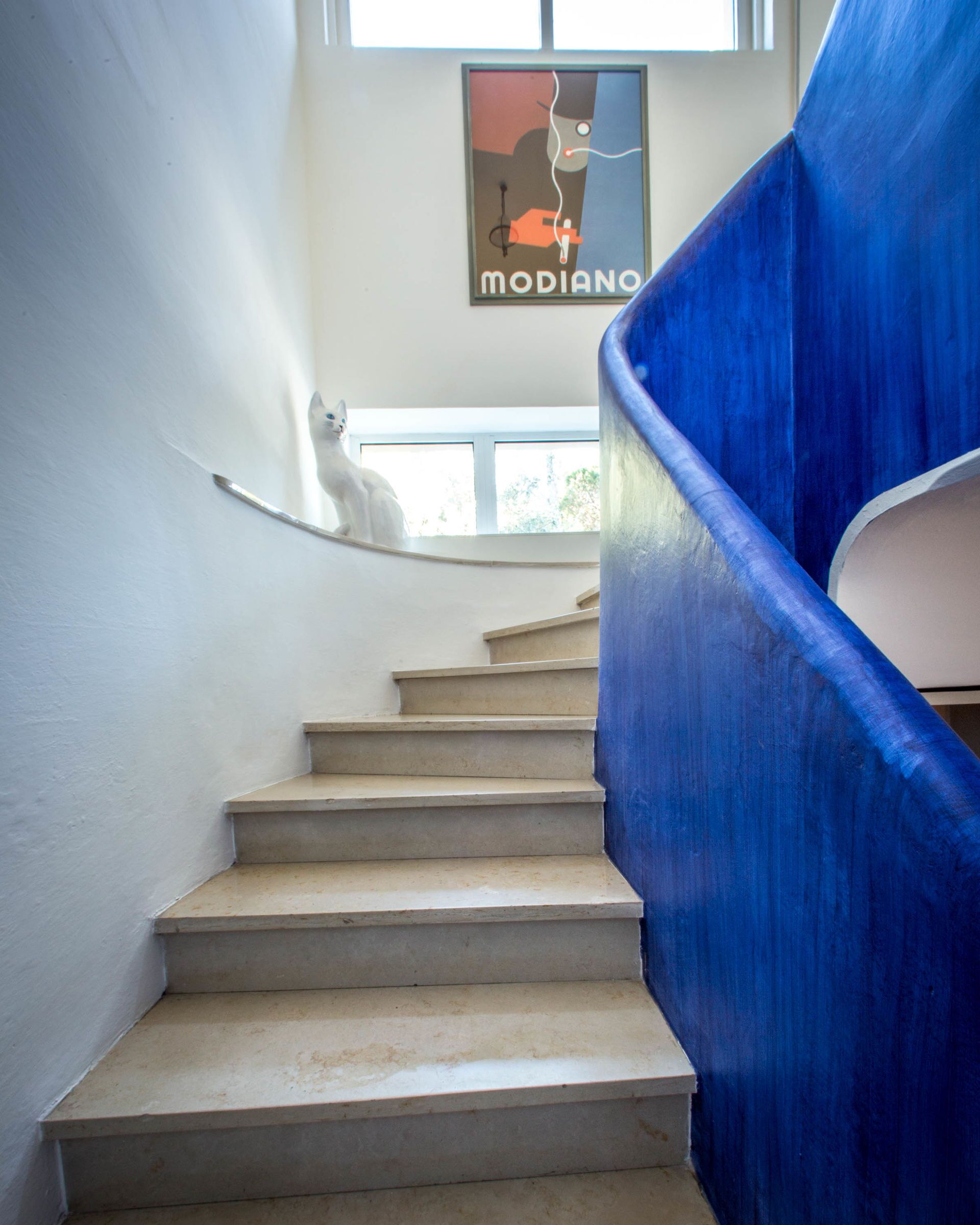 A staircase with a blue handrail, a white cat on the landing, and a poster on the wall.