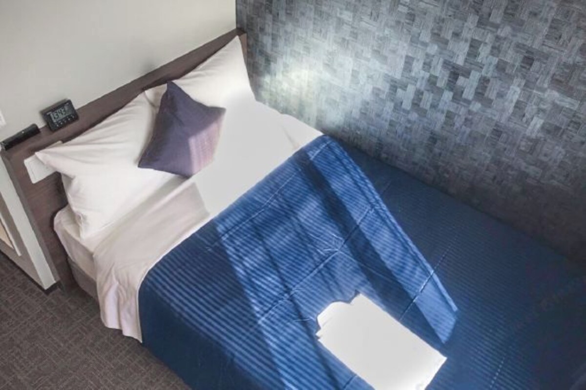 A neatly made bed with a blue and white bedspread, a pillow, and a small clock on the nightstand.