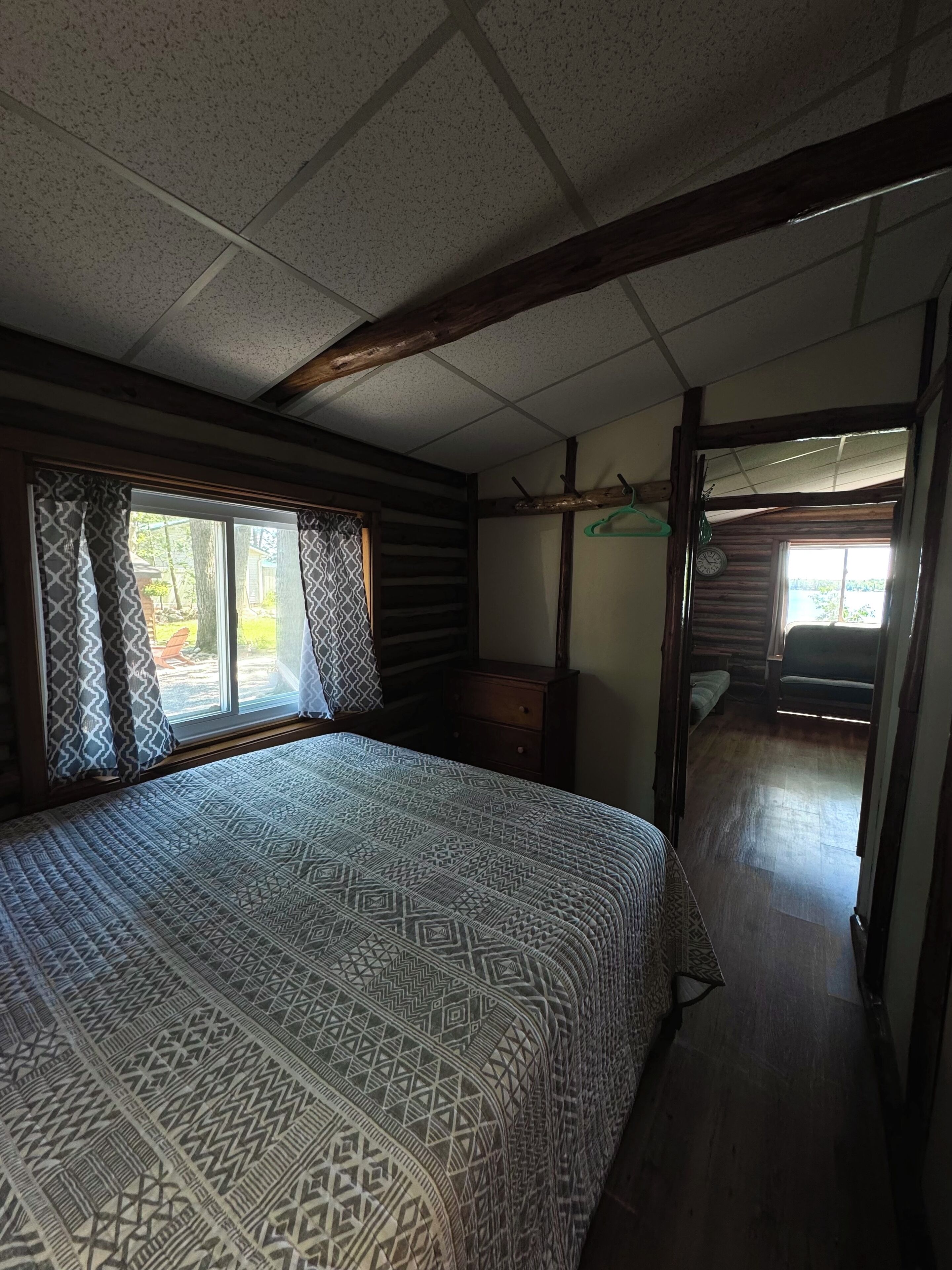 A bedroom with a large bed, wooden beams, a dresser, and a window with patterned curtains.