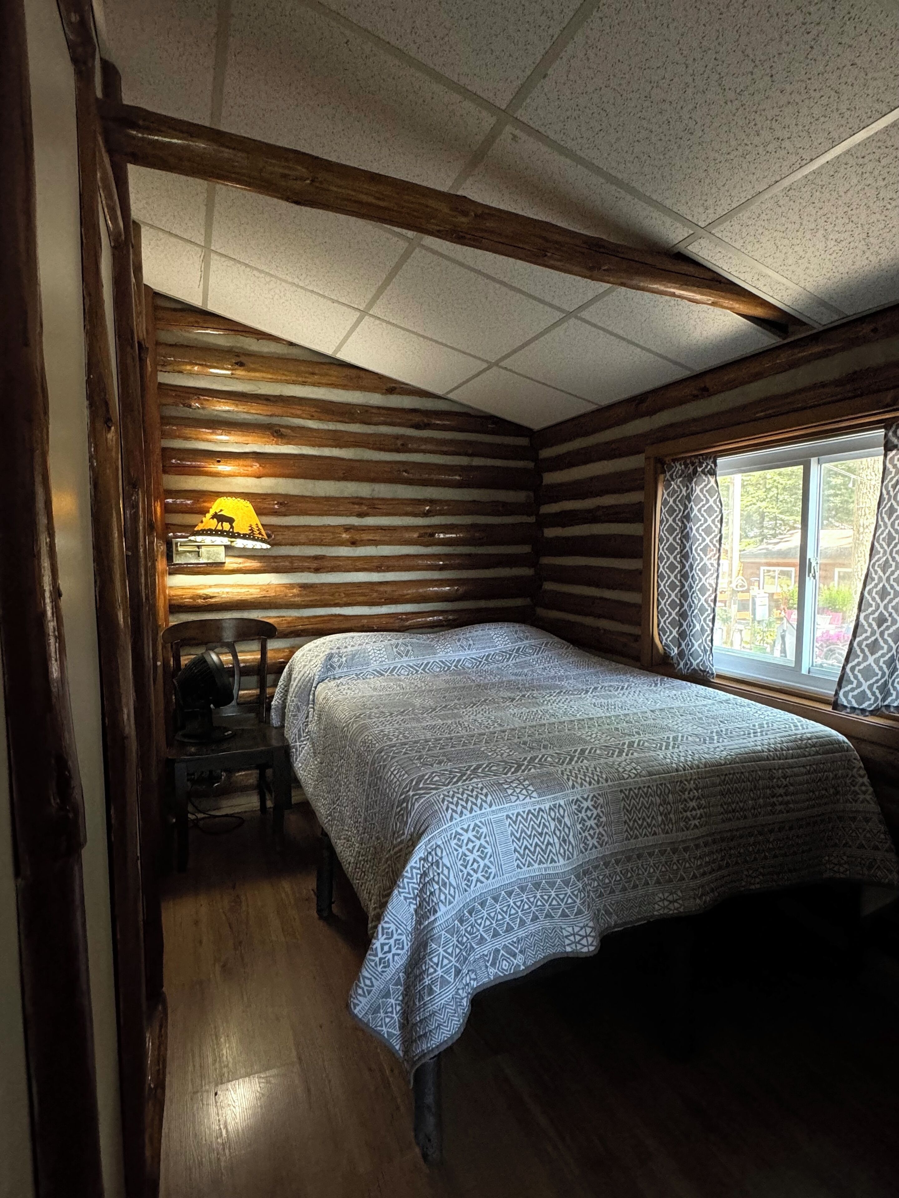 Ein Zimmer in einer Blockhütte mit einem Bett, einer Lampe und einem Fenster mit Vorhängen.