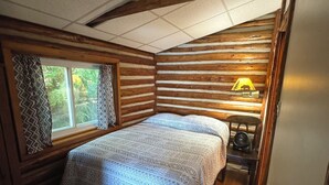 A cozy cabin-style room with a bed, wooden walls, a window with curtains, and a wall-mounted lamp. - Lake Cabin Resort - Cabin 1       (Fife Lake)