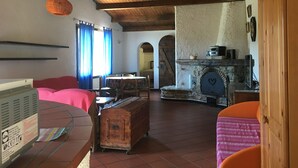 A rustic room with a wooden ceiling, a stone fireplace, a dining table with chairs, and a bed with a pink blanket.