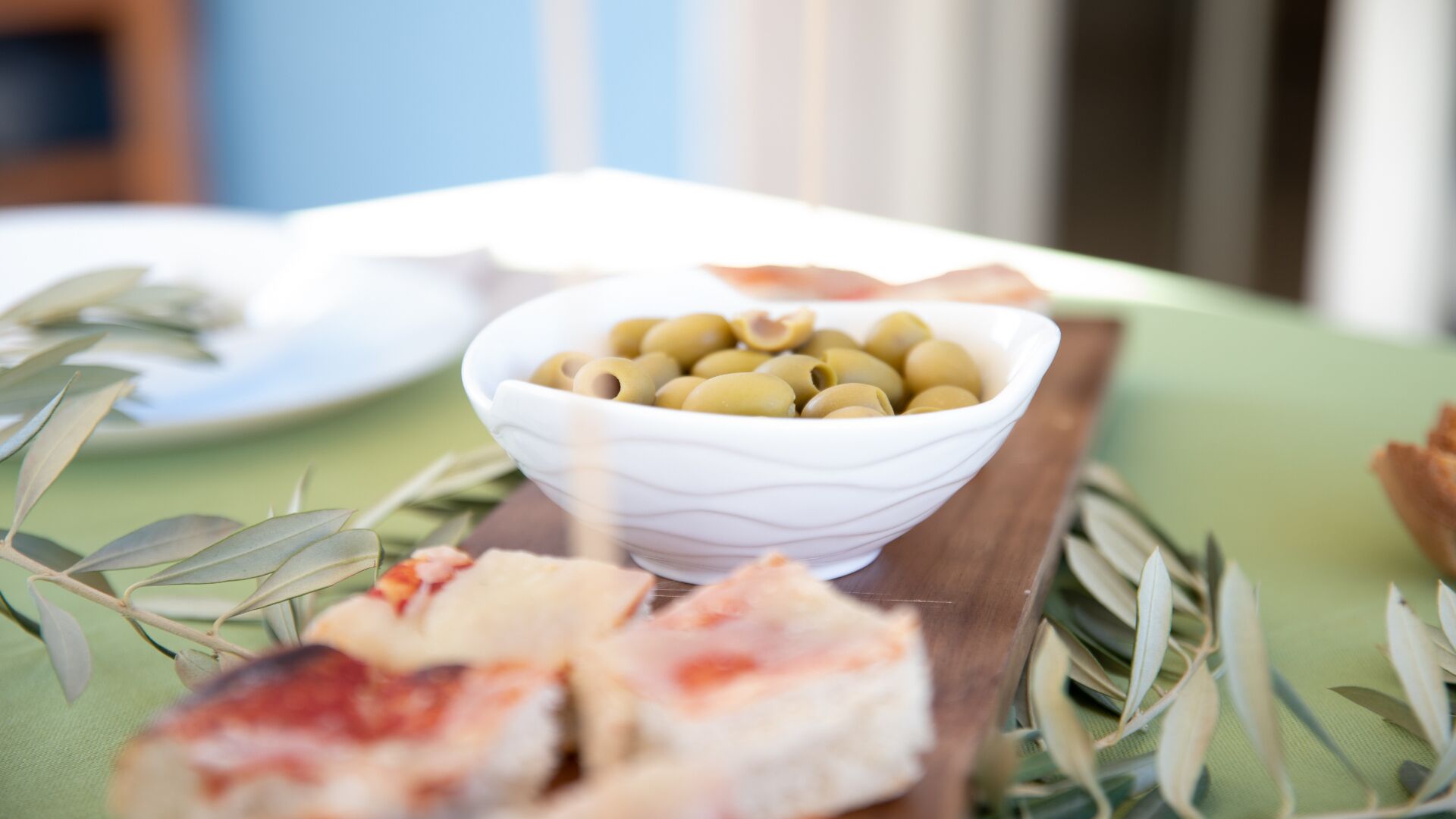 A table with a white bowl of olives, a plate with a piece of food, and a wooden board with a pastry.