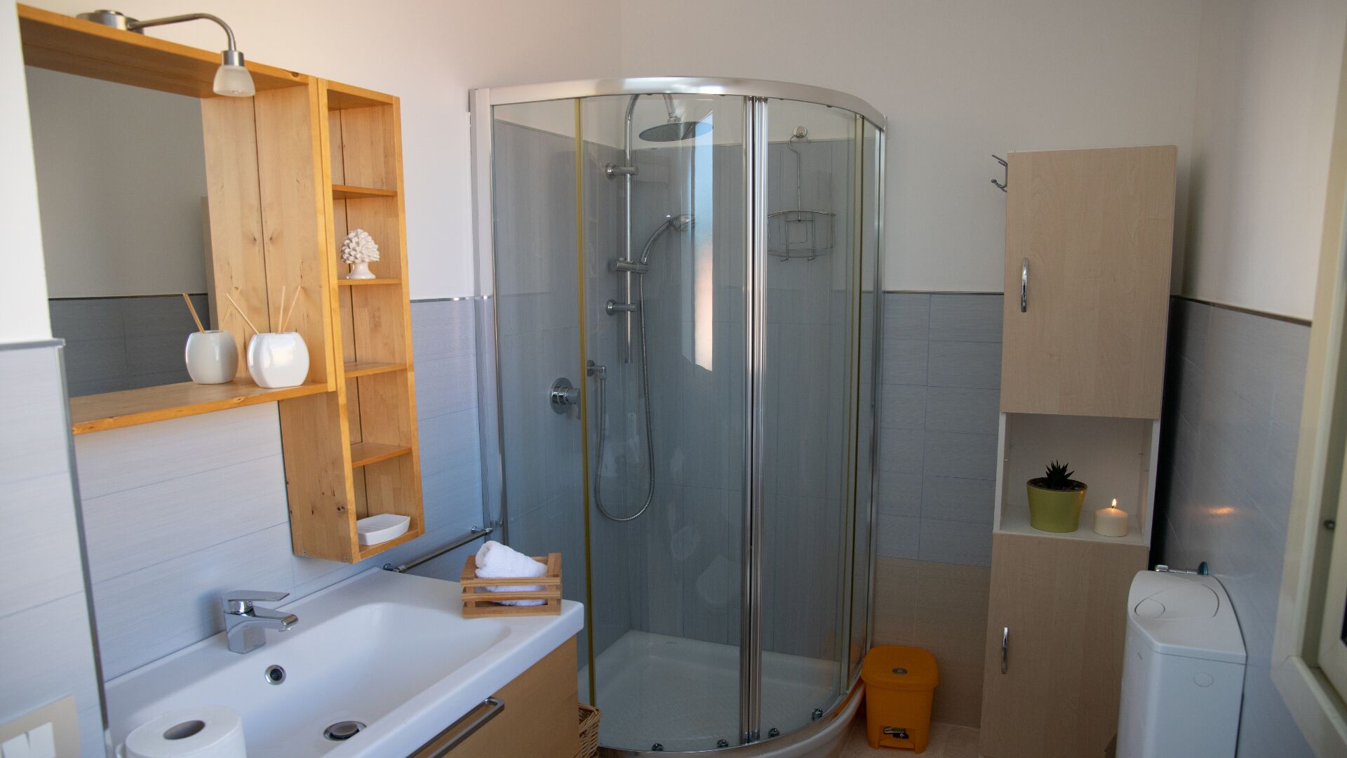 Un baño moderno con ducha envuelta en vidrio, un lavamanos blanco y un estante de madera con objetos decorativos.