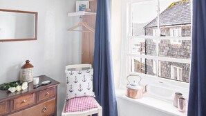 A room with a white dresser, a chair with a checkered cushion, a window with blue curtains, and a view of houses outside. - Eventide - Three Bedroom Cottage, Sleeps 5 (Kingsand)