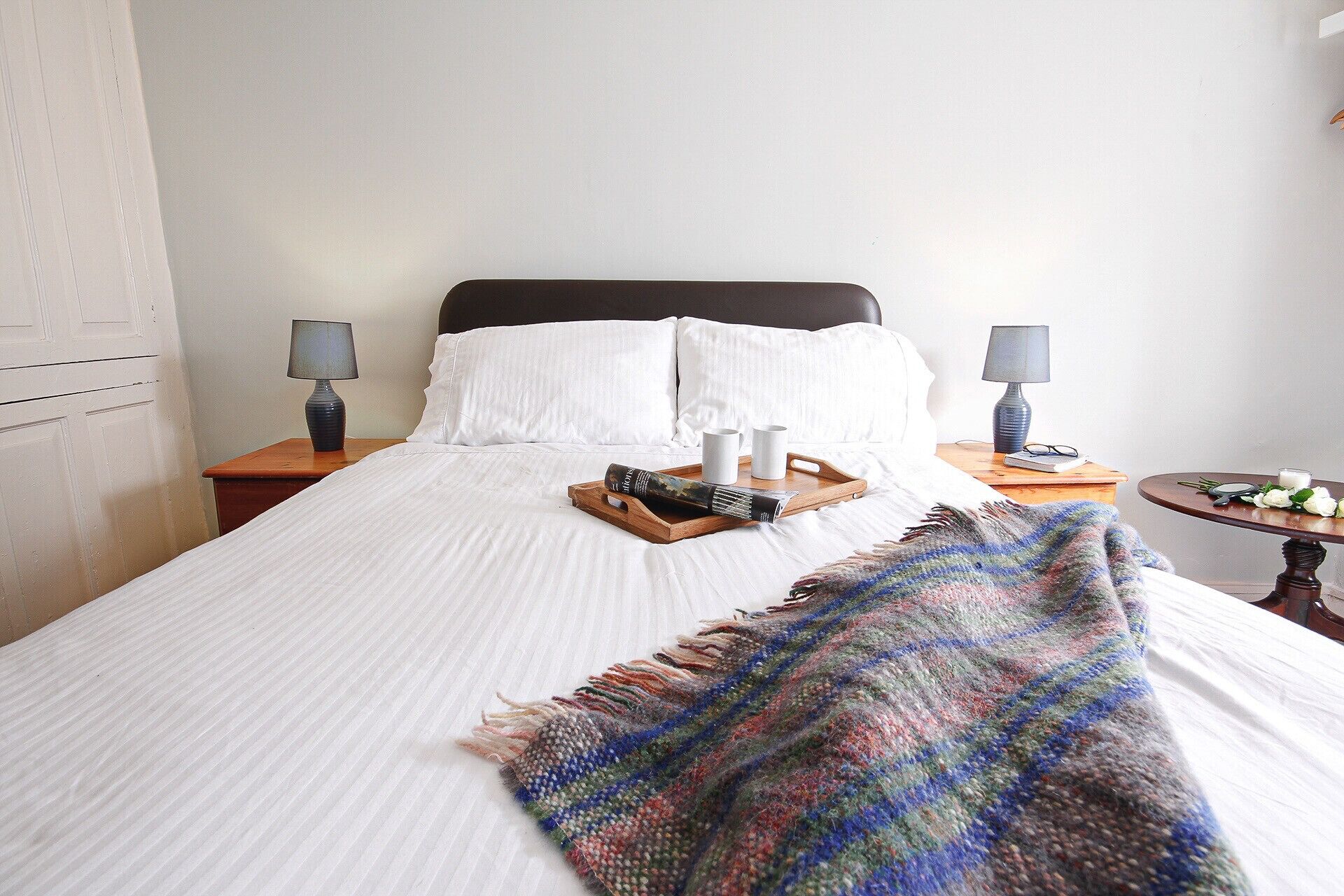 A neatly made bed with white linens, a colorful throw blanket, and a tray with a magazine and two cups. There are two bedside lamps on wooden nightstands.