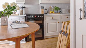 A kitchen with white cabinets, a stove, and a dining table with a plant. - Eventide - Three Bedroom Cottage, Sleeps 5 (Kingsand)