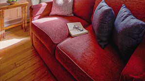 A living room with a red sofa, a wooden table with a bowl of fruit, and a view of the sea through a large window. - Eventide - Three Bedroom Cottage, Sleeps 5 (Kingsand)