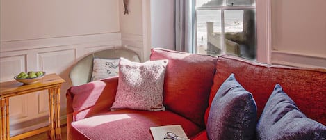 En stue med en rød sofa, et træbord med en skål frugt og havudsigt gennem et stort vindue.