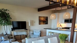 Una sala de estar moderna con comedor, que presenta una gran mesa de madera, sillas blancas tapizadas, un televisor de pantalla plana y una cocina al fondo.