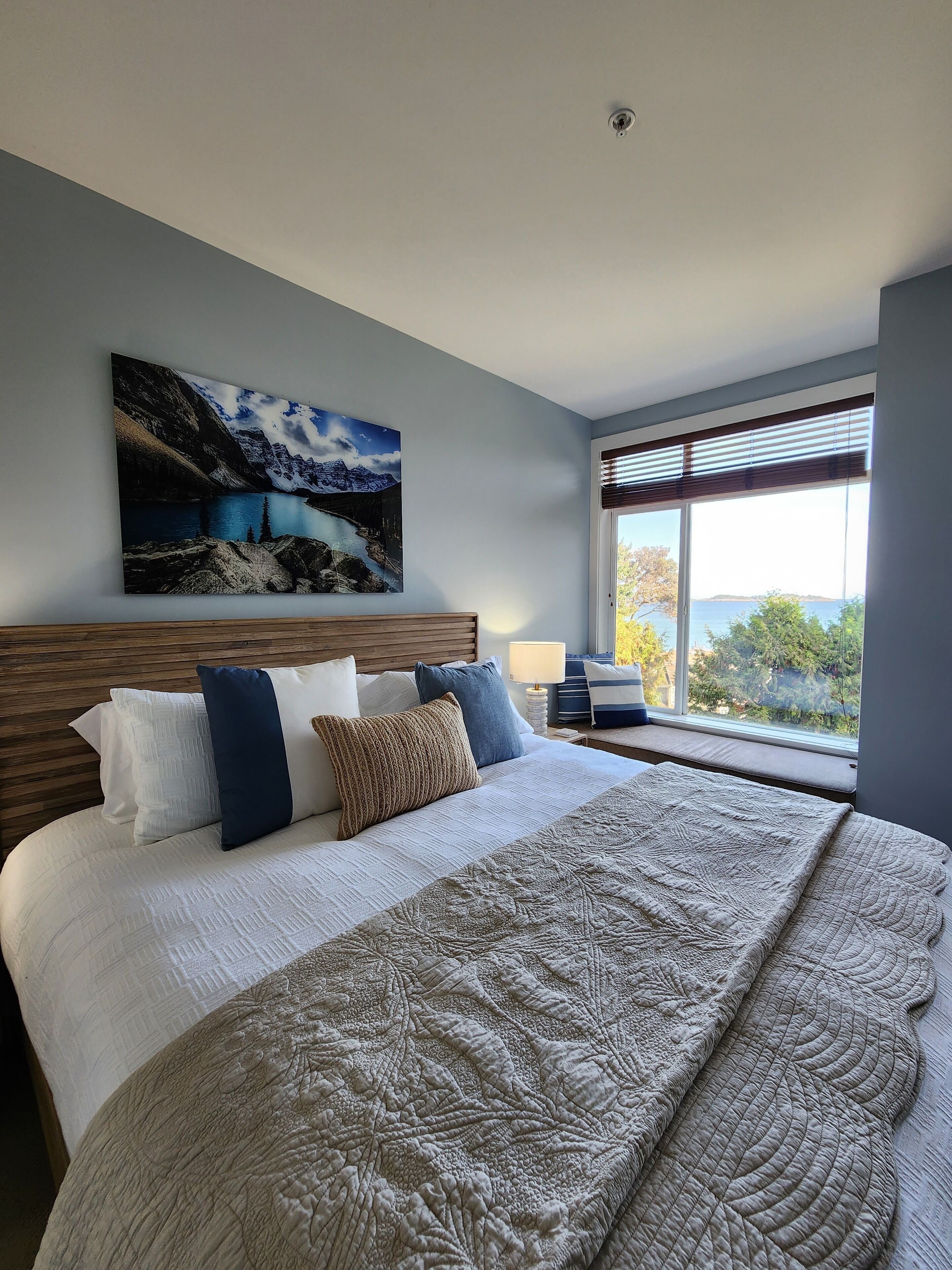 A bedroom with a large bed, a headboard, a painting of a mountain landscape, a window with blinds, and a view of trees and water.