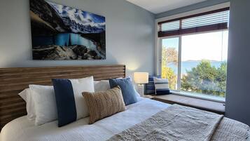 A bedroom with a large bed, a headboard, a painting of a mountain landscape, a window with blinds, and a view of trees and water.