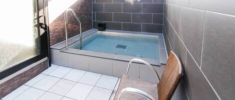 Uma pequena piscina interior com parede de vidro e escada de metal, uma cadeira e chão revestido com azulejos.