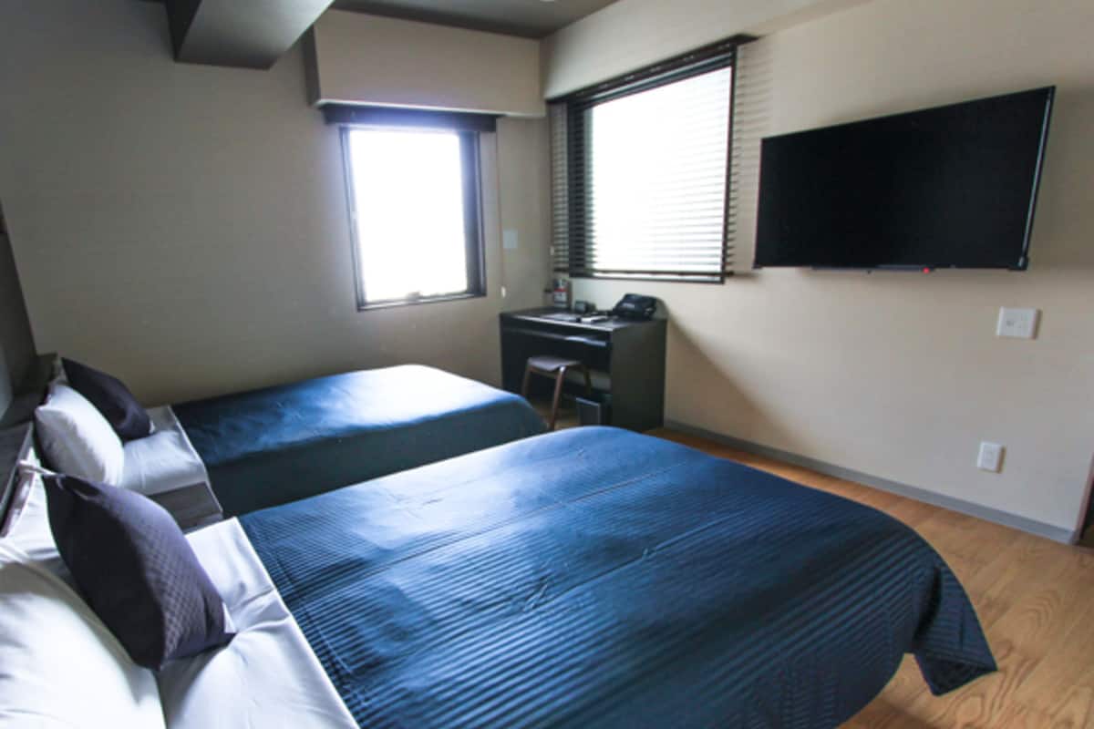 A room with two beds, a desk, a chair, a TV mounted on the wall, and a window with blinds.