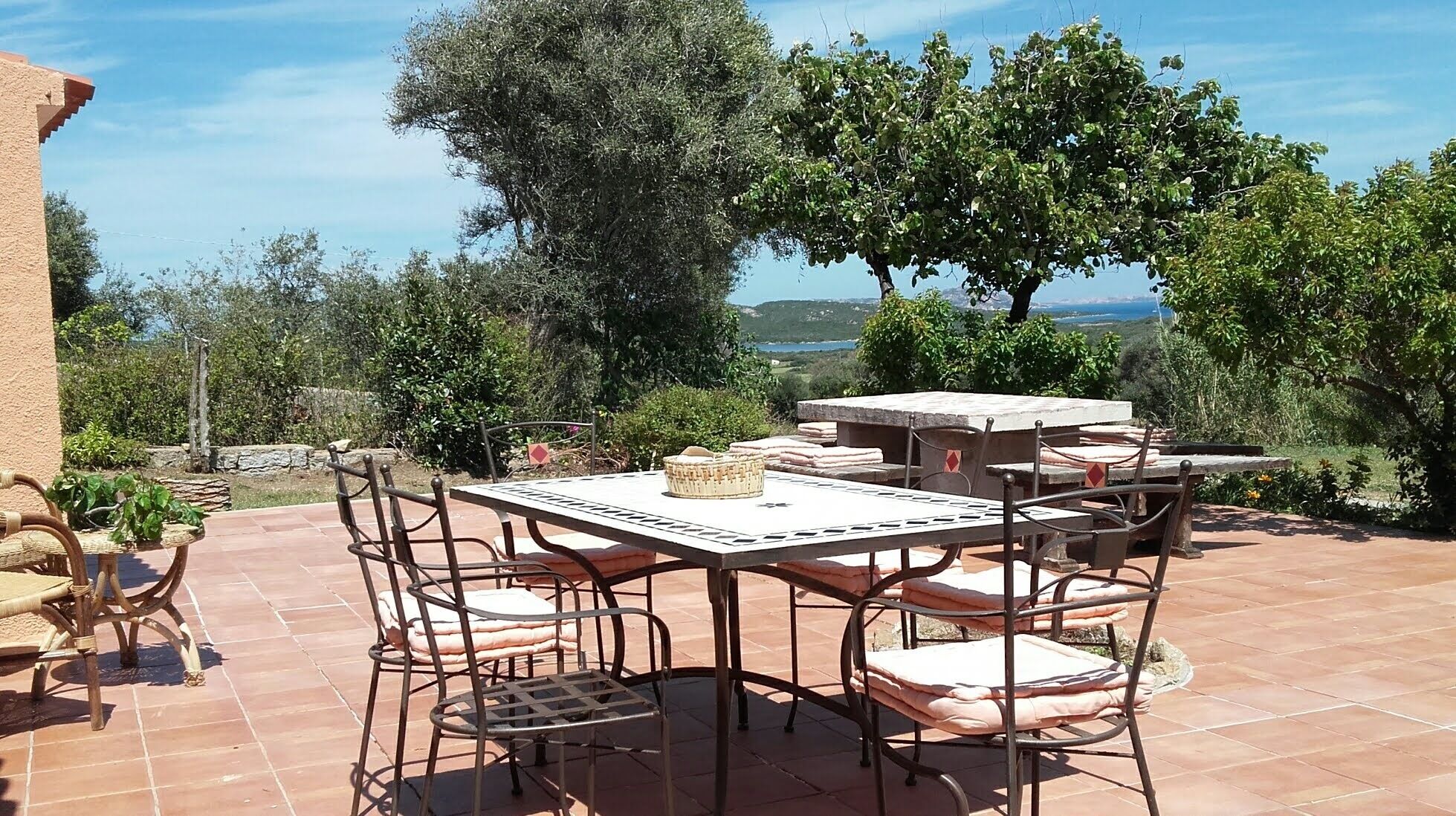 Een terras buiten met een ronde tafel en zes stoelen, omgeven door een stenen muur en bomen.