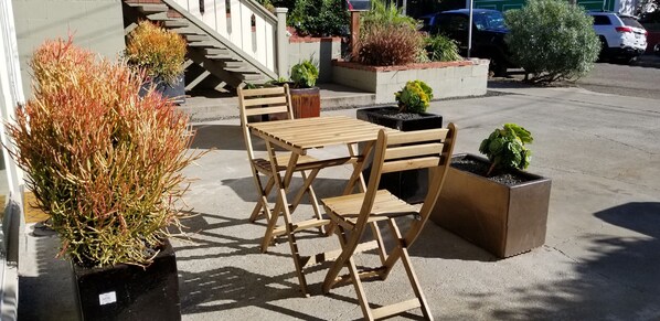 An outdoor seating area with wooden tables and chairs, surrounded by potted plants and a staircase.