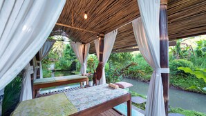 A wooden bed with a patterned bedspread, white curtains, and a view of a lush garden and water feature. - Villa Uma Anyar Ubud (Ubud)