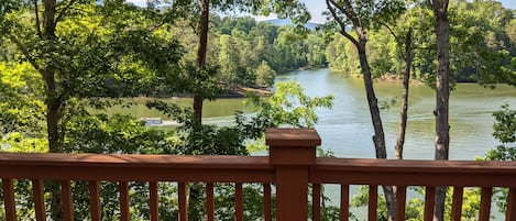 A river view from a wooden deck with a railing, surrounded by trees and a boat on the water.