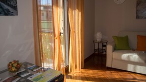A living room with a sofa, a table with magazines, a clock on the wall, and a window with sheer curtains.