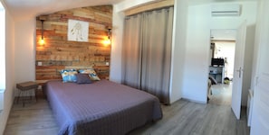 Quarto com cabeceira de madeira, cama com colcha roxa, mesa de cabeceira e luminária fixada na parede.