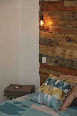 Quarto com cabeceira de madeira, cama com roupa de cama estampada, mesa de cabeceira com uma caneta e lâmpada pendente.