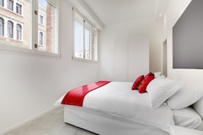 Een moderne slaapkamer met een groot bed, witte muren en uitzicht op een gebouw door een raam.