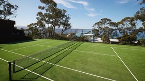 A tennis court with a net, surrounded by trees and a view of the sea.