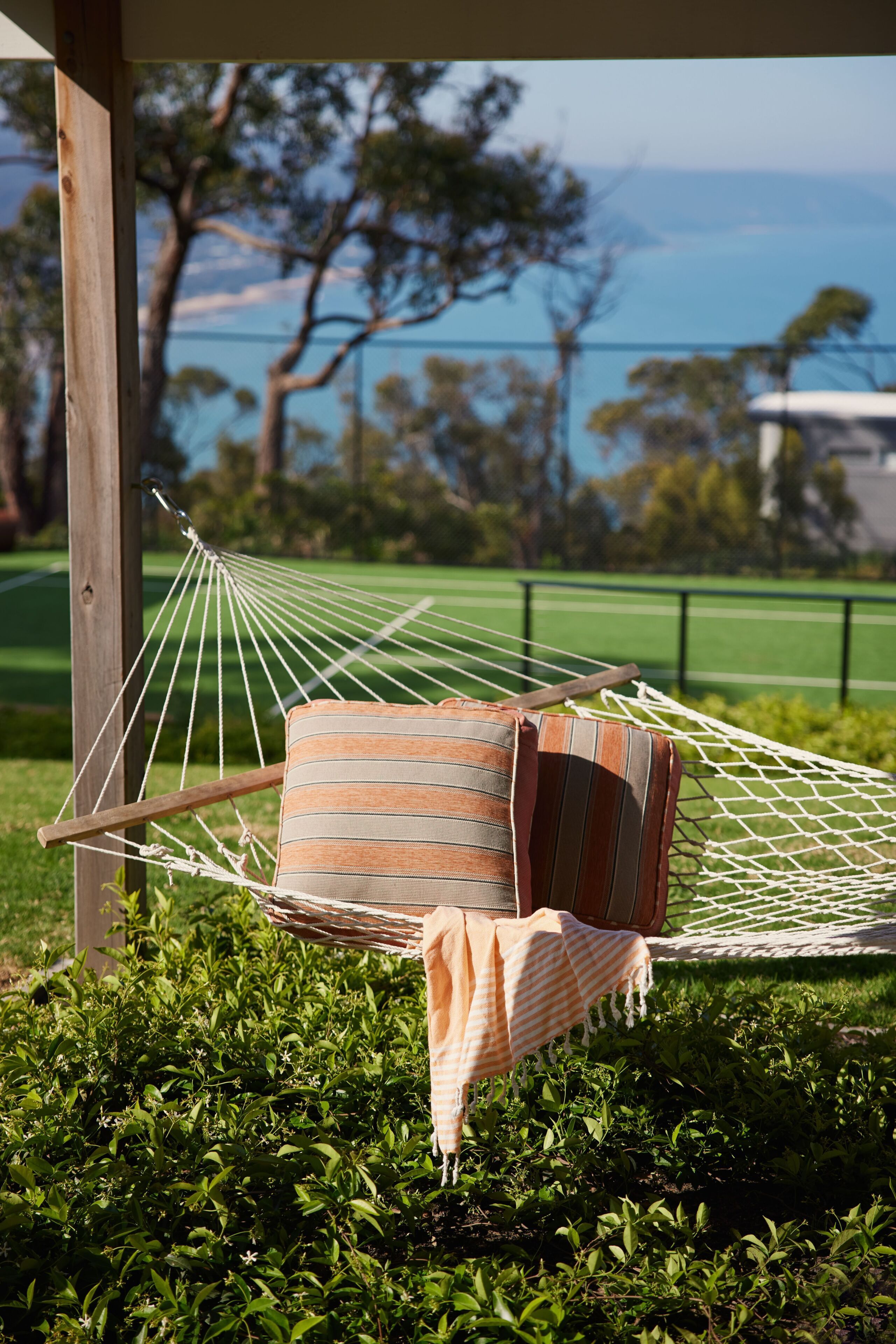 A hammock with a striped cushion and a folded blanket, set against a backdrop of greenery and a view of the sea.