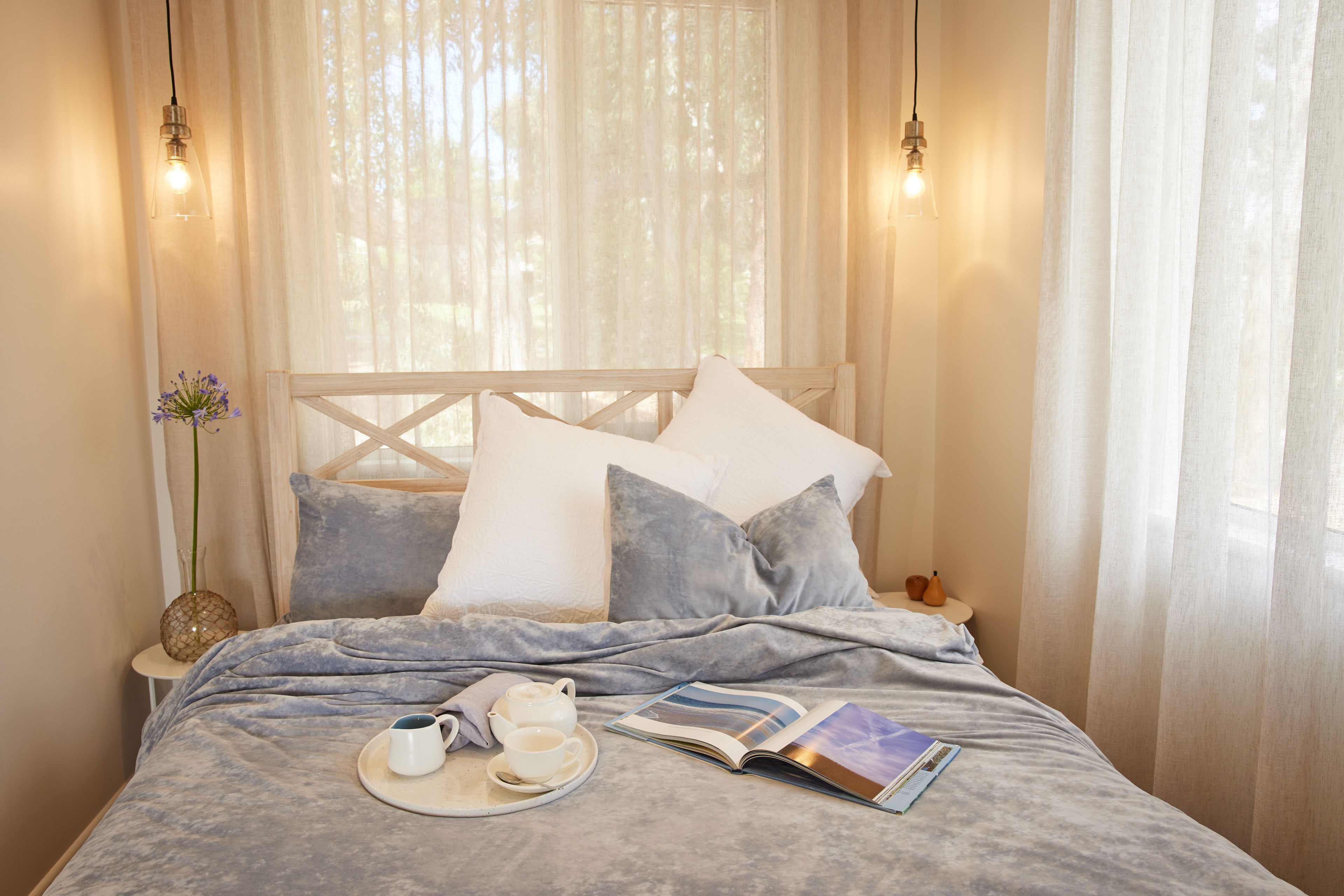 A bed with a headboard, pillows, a bedside table with a vase and a book, and two hanging light fixtures.