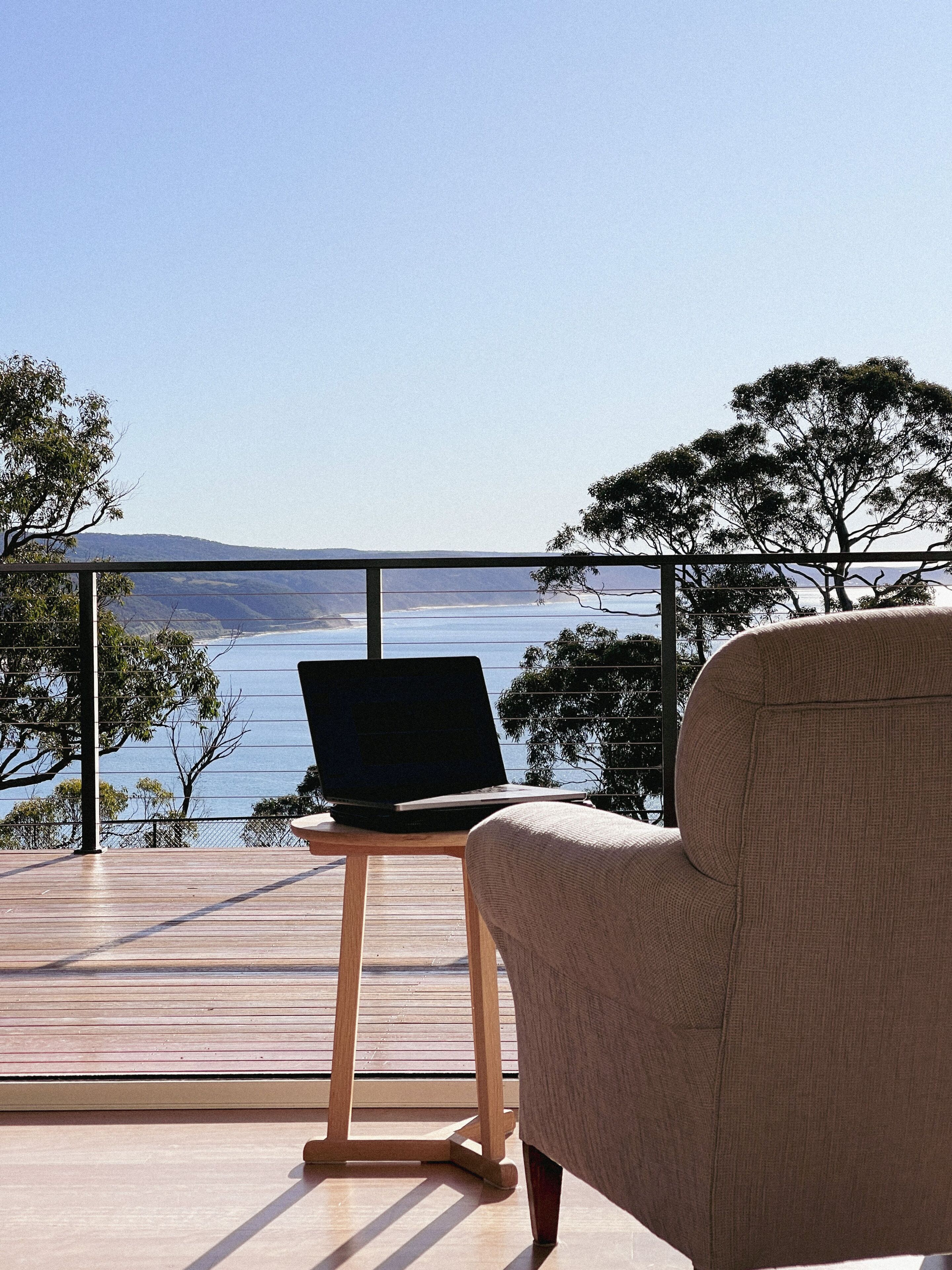 A balcony with a laptop on a wooden stand, overlooking a body of water and distant hills.