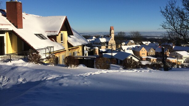 Ein schneebedecktes Dorf mit traditioneller Architektur, darunter eine Kirche mit hohem Turm und ein Wohngebiet mit Satteldächern.