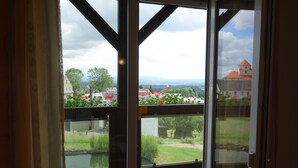 Blick von einem Balkon mit Holzboden, einer Glastür und einem Garten mit Blumen sowie einem entfernten Gebäude.