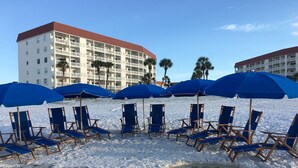 Een strand met blauwe parasols en ligbedden, een hotelgebouw op de achtergrond en palmbomen.